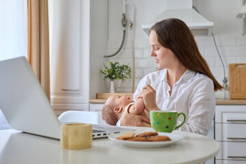 Woman sitting in kitchen with her laptop working remotely holding her newborn baby and calms down her infant using technology to balance family and business responsibilities at home