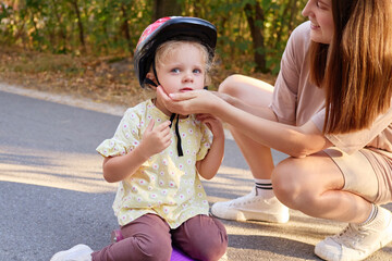 Woman helping her daughter ride skateboard in park ensuring safety with helmet before learning and...