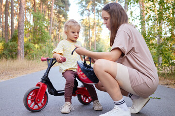 Toddler girl practicing cycling with her mother assisting her putting protective helmet during outdoor activities in park