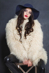 Cheerful fashion model woman in blue classic hat and white fur coat posing indoor, elegance fashion...