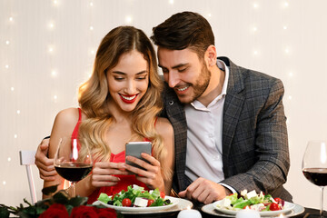 Young Couple Using Smartphone Together During Romantic Dinner In Restaurant, Browsing Internet, Social Networks Or Watching Photos. Free Space