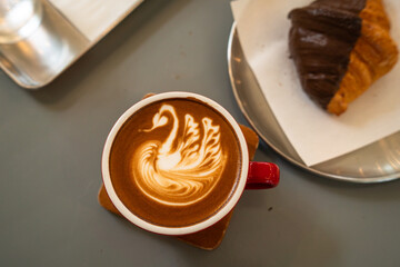Hot coffee latte with latte art milk foam a swan shape in cup mug with chocolate Croissant on wood desk on top view.As breakfast In a coffee shop at the cafe,during business work concept,Rainy season © Thinapob