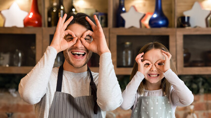 Fooling Around Together. Happy Dad And Daughter In Aprons Making Glasses With Hands In Kitchen, Having Fun At Home, Posing To Camera