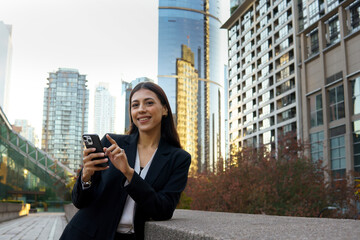 Fototapeta premium Attractive Hispanic businesswoman using mobile phone outdoor with background of modern glass buildings