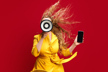 Obraz premium Young woman in red dress announcing sale season in megaphone, holding mobile phone against red studio background. Online shopping. Concept of sales, shopping, news, announcement, advertisement