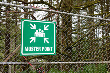 A green muster point safety sign displayed on a chain-link fence in a forested area, indicating an...