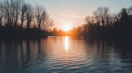 A tranquil lake with bare trees silhouetted against a vibrant sunset. The sun's golden rays create a shimmering reflection on the calm water, evoking a sense of peace and serenity.