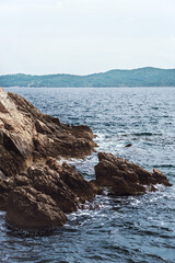 A rocky coastline with jagged stone formations extending into the sea. The waves gently crash against the rocks. The calm, blue ocean, where distant, low-lying hills are visible under a light. 