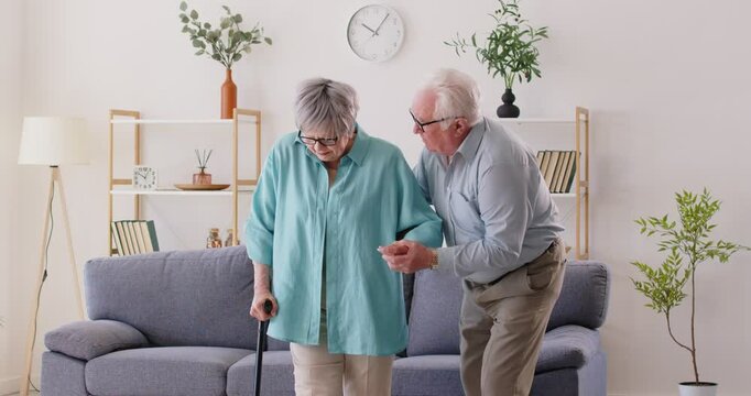 Elderly man is gently helping his wife as she walks with aid of cane in their living room. Concept of family relationships, elderly couple, care, health and illness in old age. 4k video footage.