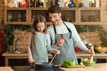 Young man and his little daughter cooking together in kitchen, preparing healthy meals with frying pan, enjoying homemade food, making vegetable salad for lunch, having fun at home, free space