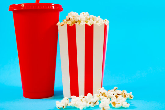 Horizontal image of red cardboard bucket with white stripes with popcorn and red soda glass on a blue background, concept of cinema food and entertainment