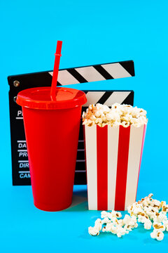 Vertical image of movie clapperboard behind red cardboard bucket with white stripes with popcorn and red soda cup on blue background, concept of food and entertainment in cinema