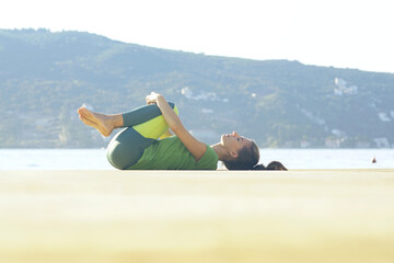 Girl in janyasana pose lying on the beach with sea and hill in the background. Yoga classes in picturesque places outdoors: young woman doing morning workout alone on sunny warm day.