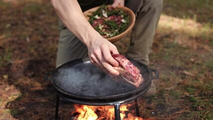 Close-up of a person's hands placing seasoned meat into a hot, smoking cast iron pan over a campfire. The meat sizzles as it makes contact with the pan, surrounded by flames.