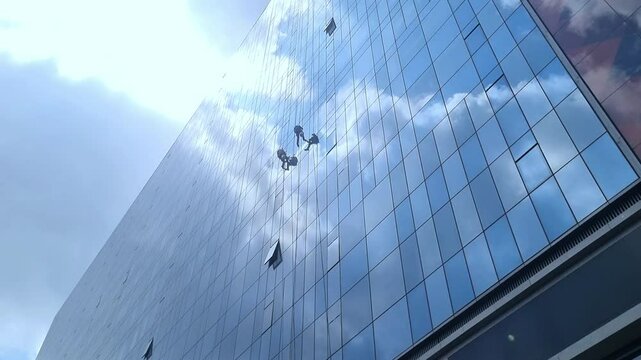 Window Cleaners Working on Glass High-Rise Building, Window cleaners suspended on a skyscraper, methodically cleaning the reflective glass facade of a modern high-rise building.