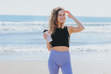 Fitness, sport and healthy active lifestyle. Happy slim young athlete lady in sportswear holds bottle of water, rests after workout on sea beach. Break, hydration, wellness, healthcare, aqua balance