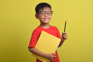 Asian school boy student wearing glasses holding books and pencil isolated on yellow background. Back to school concept