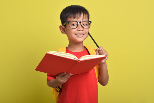 Asian boy with backpack holding pencil and open book posing thinking solving problem isolated on yellow background. Time to study. Back to school