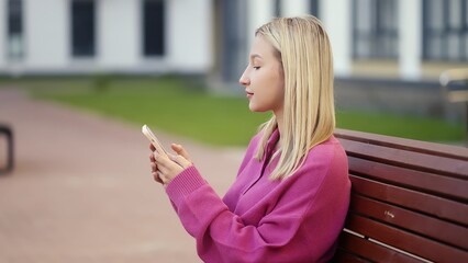In a tranquil outdoor environment, a young woman sits comfortably on a bench, engrossed in her...