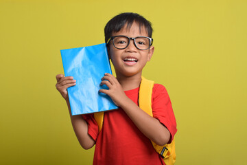Asian boy holding blue book in his hand isolated on yellow background. A little boy excited for school