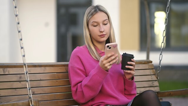 On a delightful summer day, a trendy young woman finds joy in using her phone while swinging in a beautiful park