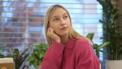 A young woman in a pink outfit is deep in thought, surrounded by lush plants and bright natural...