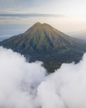 Aerial view of a majestic volcanic mountain surrounded by lush greenery and clouds at sunset, Licin, Banyuwangi, East Java, Indonesia.