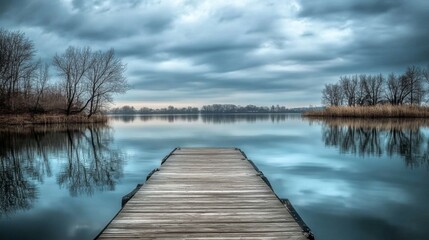 A wooden pier stretches into a calm lake, surrounded by trees with bare branches. The overcast sky casts a serene ambiance, reflecting soft hues on the water at early dawn.