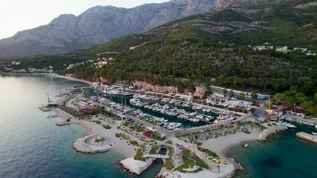 Aerial view of the picturesque harbour and village with boats, surrounded by clear turquoise sea and mountains, Baska Voda, Croatia.