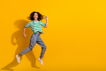 Full body portrait of nice young girl jump raise fist empty space wear striped t-shirt isolated on yellow color background