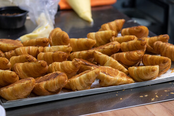 Close-up side view of fresh baked brown croissants lying upside down on baking tray before filling with cream on professional kitchen table. Soft focus. Copy space. Food preparation theme.