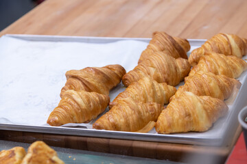 Close-up side view of fresh baked brown croissants lying on white wax paper on baking tray on professional wooden kitchen table. Soft focus. Copy space. Puff pastry preparation theme. Food background.
