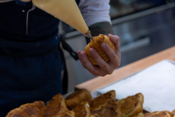 Close-up side view of cook filling fresh baked croissant with custard using icing bag in professional bakery kitchen. Soft focus. Pastry preparation  theme.