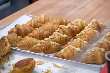 Close-up side view of fresh baked brown croissants lying on white wax paper on baking tray on professional wooden kitchen table. Part of buns covered with custard. Puff pastry preparation theme.