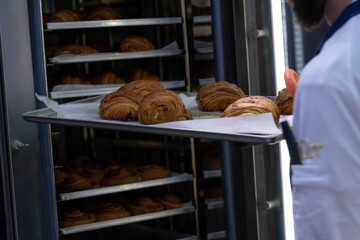 Rear view of chef taking off baking tray with fresh baked brown croissants from professional convection oven in bakery. Soft focus. Puff pastry preparation theme.