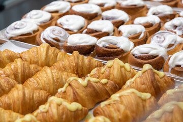 Close-up side view of various fresh baked puff pastry items (croissants with different fillings and cinnamon rolls) on baking trays on professional kitchen table. Soft focus. Food background.
