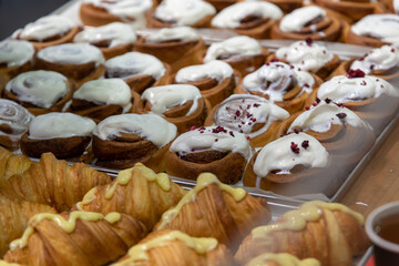 Close-up side view of various fresh baked puff pastry items (croissants with different fillings and cinnamon rolls) on baking trays on professional kitchen table. Soft focus. Food background.