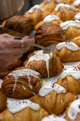 Close-up side view of baker pouring glazing on top of fresh baked brown chocolate croissants using spoon on professional kitchen table. Soft focus. Food preparation and decoration theme.