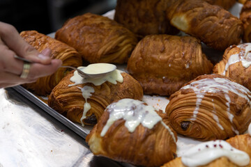 Close-up side view of baker pouring glazing on top of fresh baked brown chocolate croissants using spoon on professional kitchen table. Soft focus. Food preparation and decoration theme.