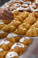 Close-up side view of person putting edible flowers using tweezers on top of fresh baked brown croissants with various fillings on kitchen table. Soft focus. Food preparation and decoration theme.