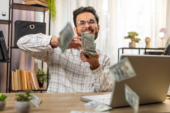 Cheerful rich Indian businessman working on laptop and wasting throwing money cash more tips earnings big profit success win lottery shopping. Happy Arabian freelancer man manager at home office desk