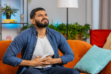 Hispanic young man sitting on sofa feeling sudden strong abdominal stomach ache, gastritis problem....