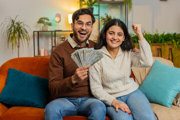 Happy young Indian couple holding showing money cash banknote together looking at camera sitting on home sofa in room. Successful rich diverse family clenching fists celebrate lottery jackpot game win