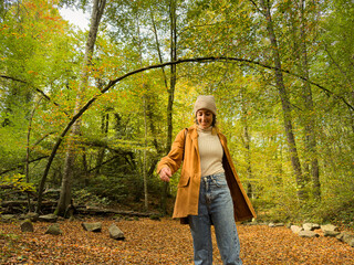 A woman is walking in a forest wearing a brown coat and jeans