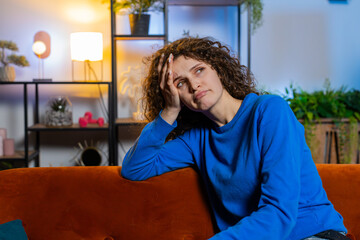 Portrait of upset lonely young woman with curly hair thinking about problems sitting on sofa in...