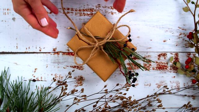 Hands tying a rustic gift with natural twine, decorated with pine sprigs and berries. Perfect for showcasing holiday gift-wrapping, DIY, and nature-inspired seasonal aesthetics. Flat Lay top view.