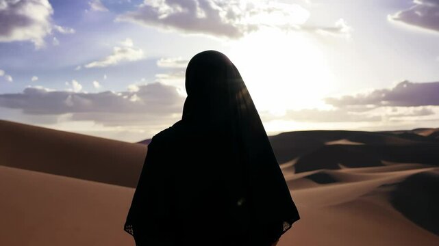 Muslim woman in hijab. A woman wearing a black head scarf stands in a desert. The sky is blue and the sun is setting
