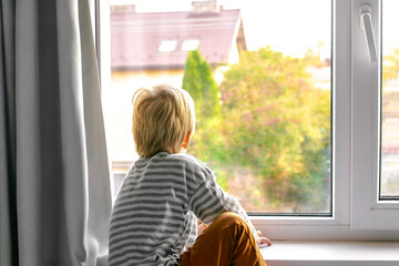 Little boy child kid in pajamas looking sadly through window and wants to go out for a walk and...