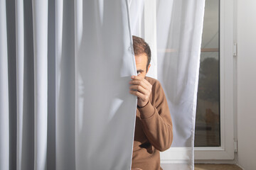 Caucasian man looks out from behind a curtain.