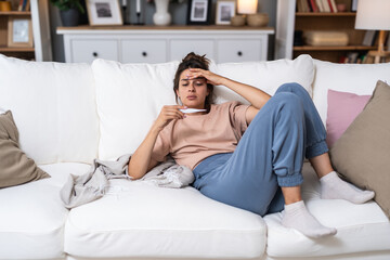 Fototapeta premium Sickness seasonal virus problem concept. Woman being sick having flu lying on sofa holding thermometer. Sick female lying in bed with high fever. Cold flu and migraine with high body temperature.